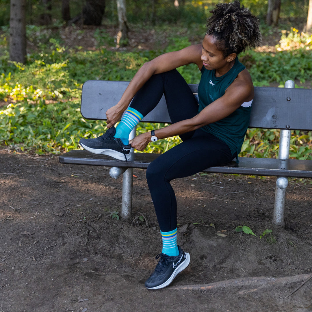 Full body shot of model sitting on a bench putting on sneakers wearing women's stride micro crew running sock in teal