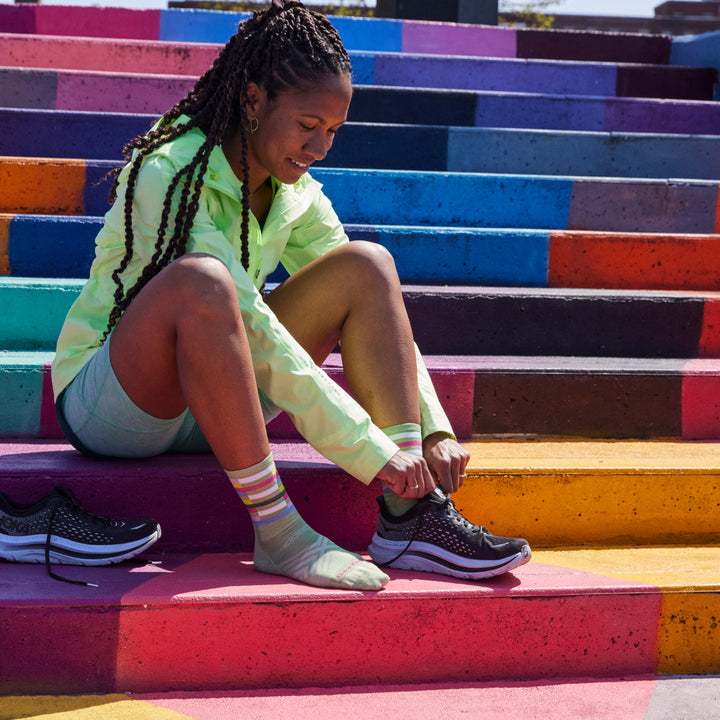 Model sitting on colorfully painted concrete steps and wearing 1063 socks in Mint colorway while lacing up black running shoe on left foot. 