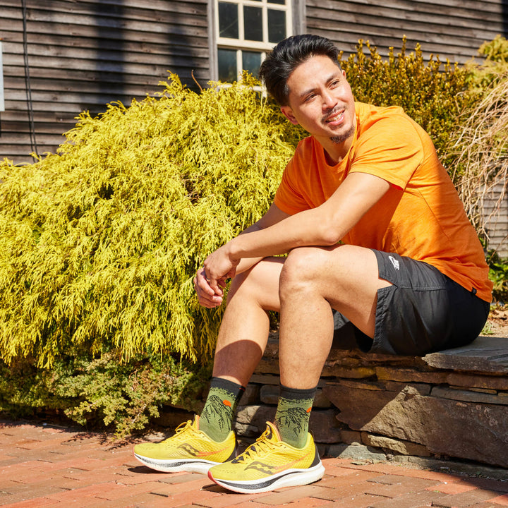 Model sitting on rock wall with sneakers on wearing the 1066 Vert Micro Crew running sock in Lichen