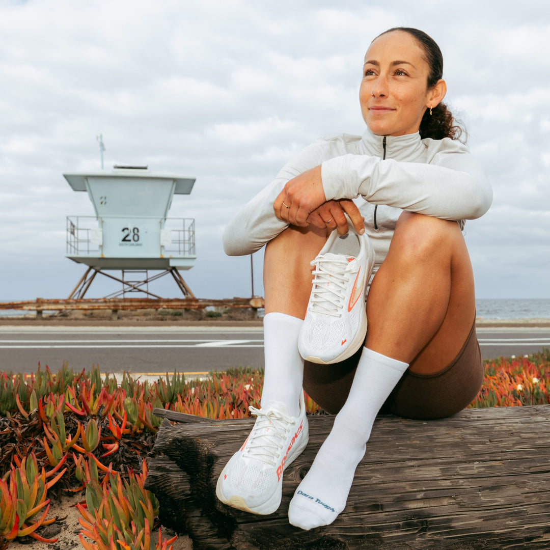 Model sitting on log in front of life guard tower holding one shoe wearing the 1068 micro crew running sock in white colorway