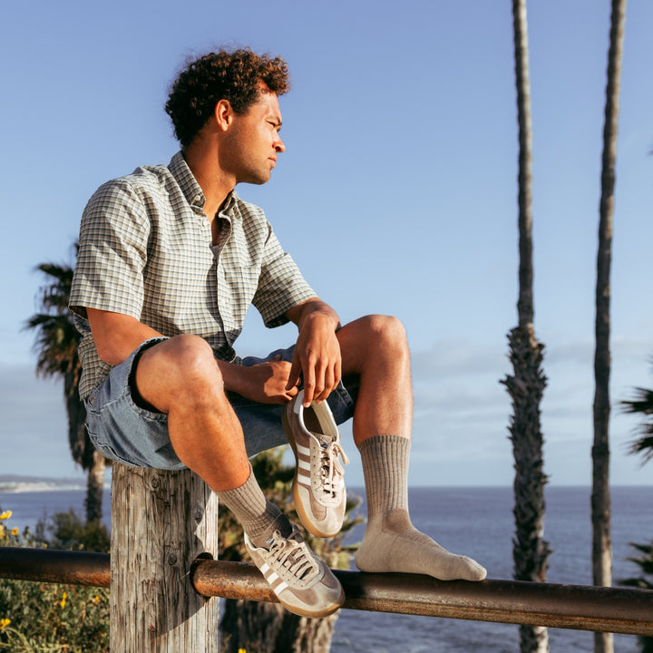 model sitting on fence poll holding one shoe wearing the 1657 standard lightweight lifestyle crew in rye colorway