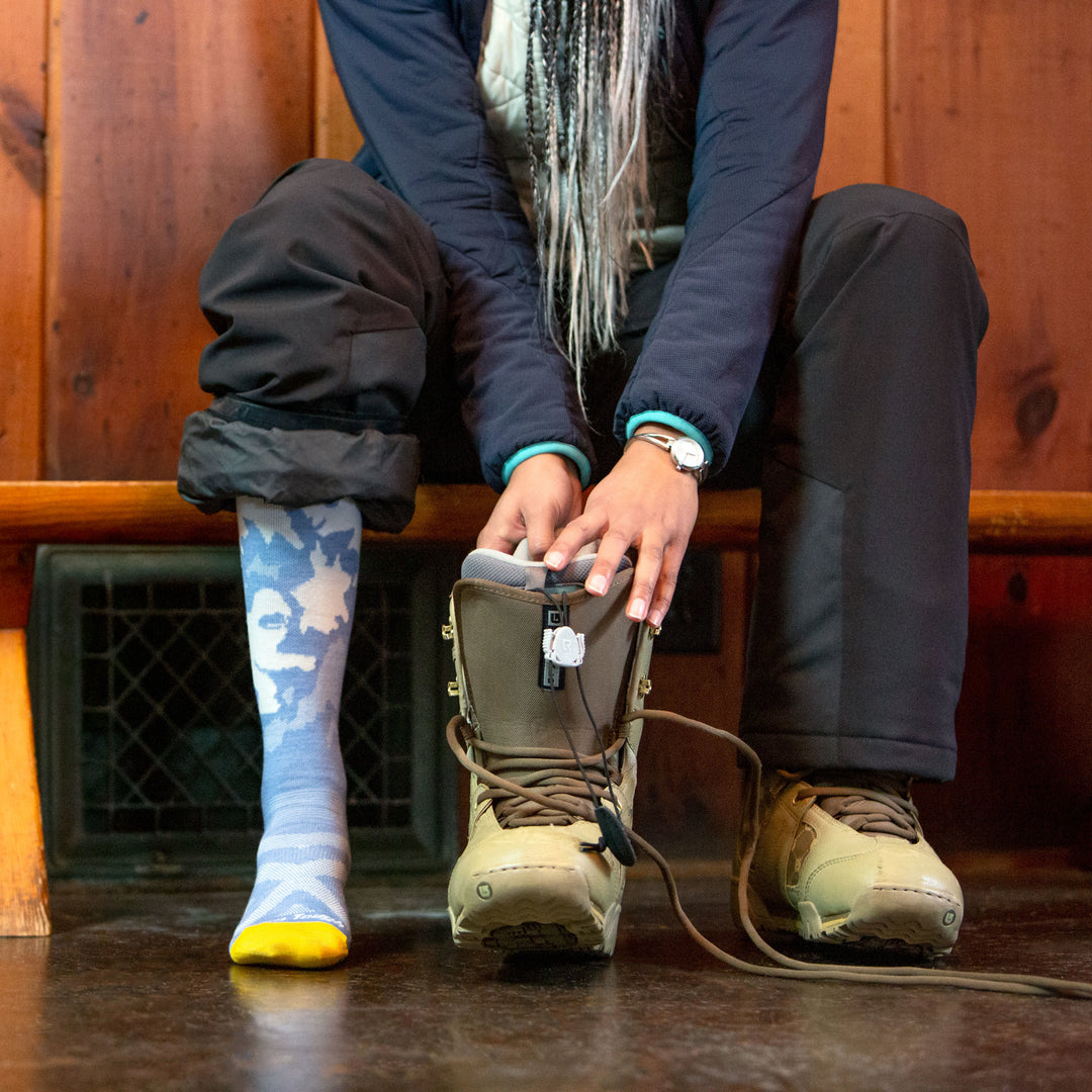 Model sitting inside wearing women's yeti over-the-calf ski sock in midnight blue while putting on snowboard boots