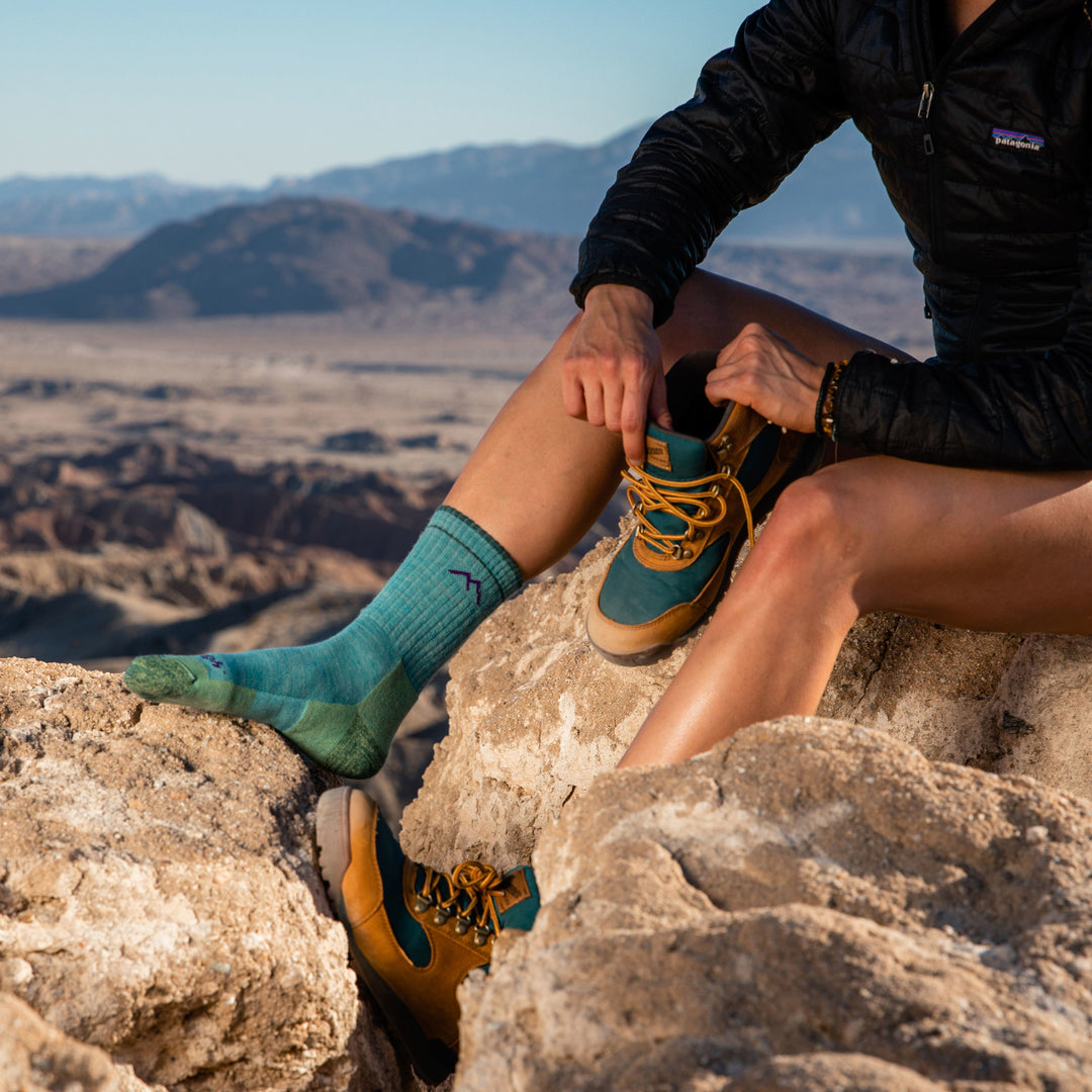 Side shot of model sitting on rock wearing women's hiker micro crew hiking sock in aqua while putting on gold hiking shoes