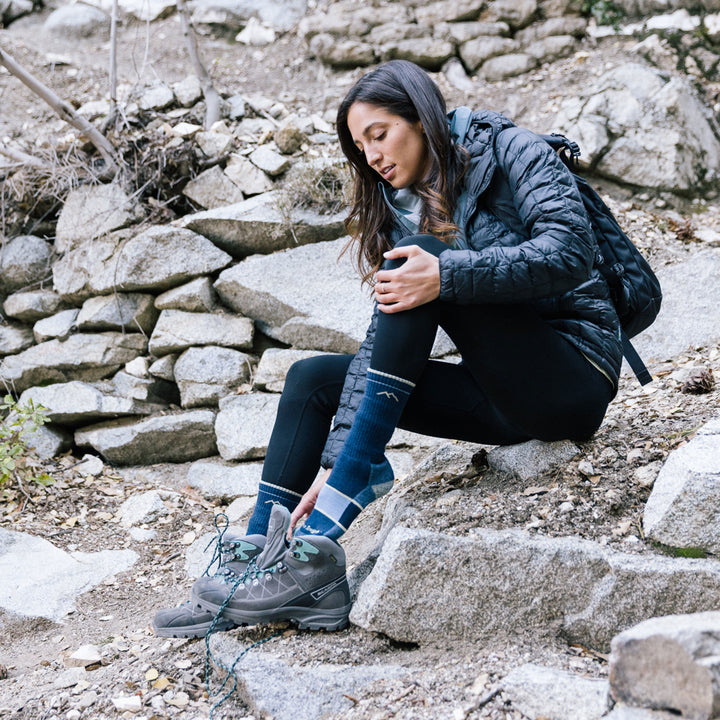 Model sitting on rocks wearing women's hiker boot hiking sock in navy eclipse while putting on gray hiking shoes
