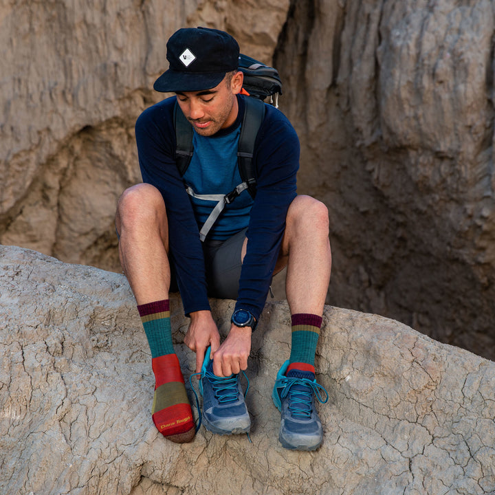 Model sitting on a rock wearing men's heady stripe miro crew hiking sock in teal while putting on blue shoes