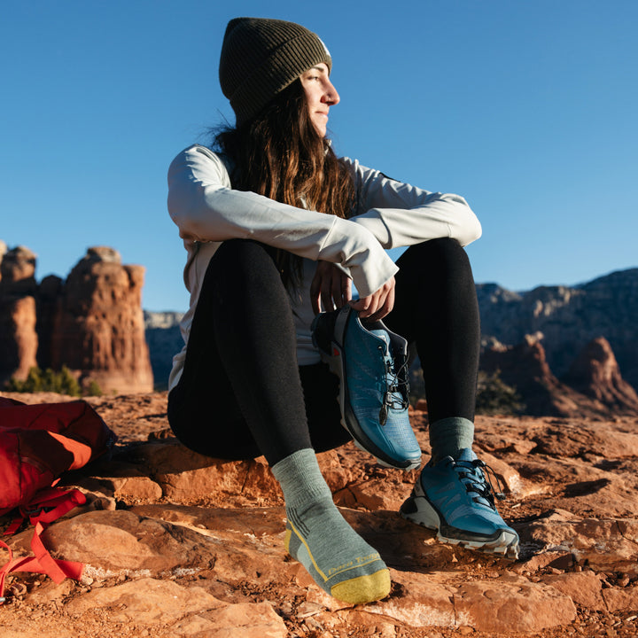 Image of model sitting on red desert rocks wearing 1958 socks in Sage colorway while wearing one blue hiking shoe and holding the other