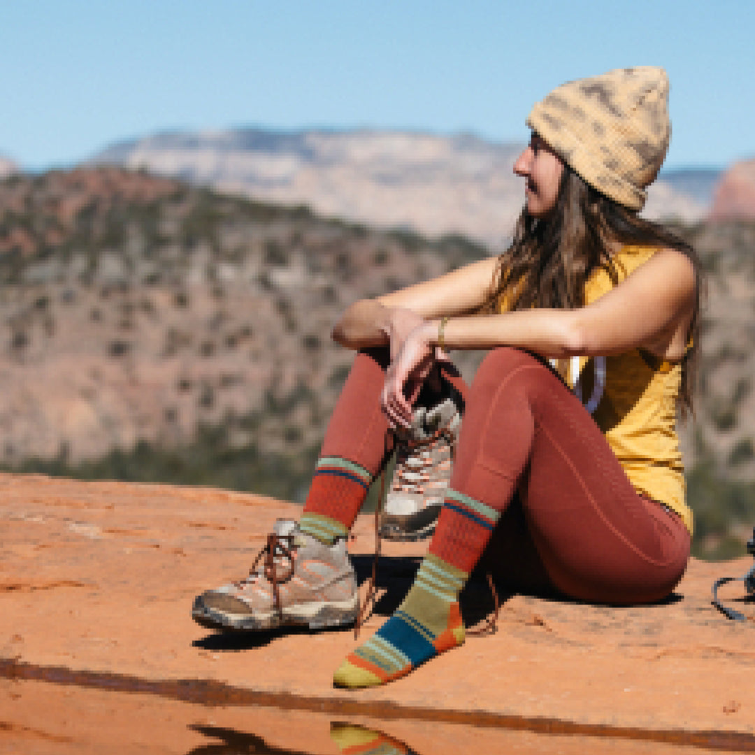 Image of model sitting on red desert rocks wearing 1969 socks in Sandstone colorway while wearing one brown hiking boot and holding the other