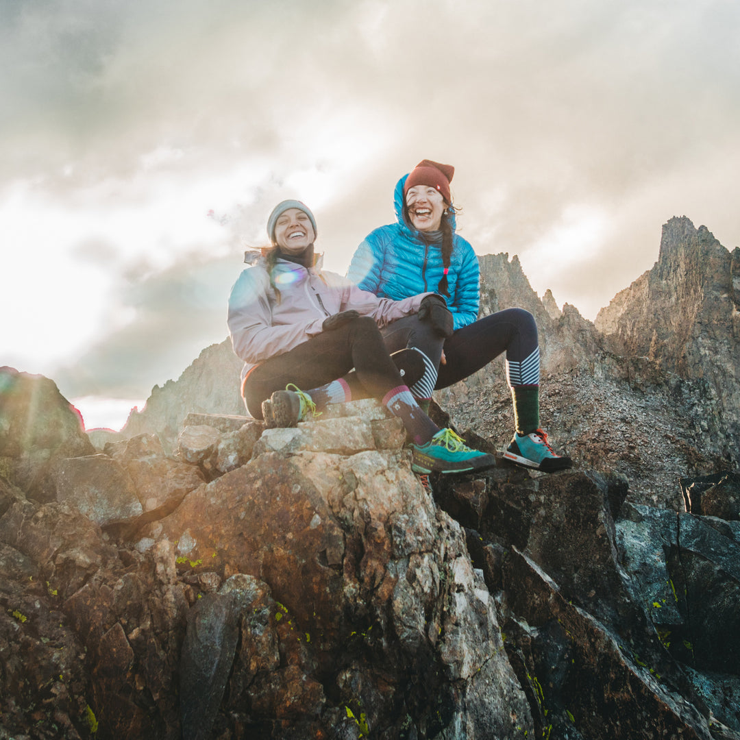 Image of 2 women, laughing, sitting on a rocky mountaintop and one is wearing Women's Bear Town Hiking sock in Purple