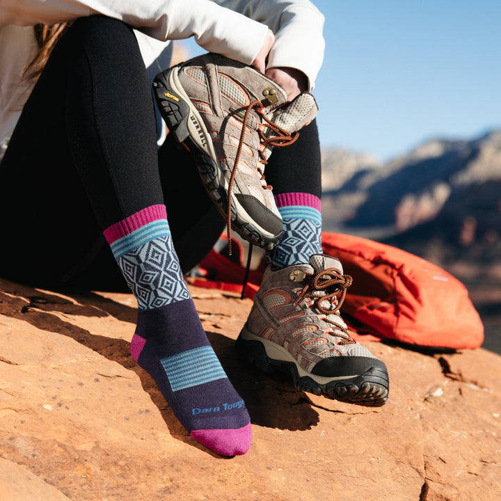 Image of model sitting on red desert rocks wearing 1977 socks in Blackberry colorway while wearing one gray hiking boot and holding the other