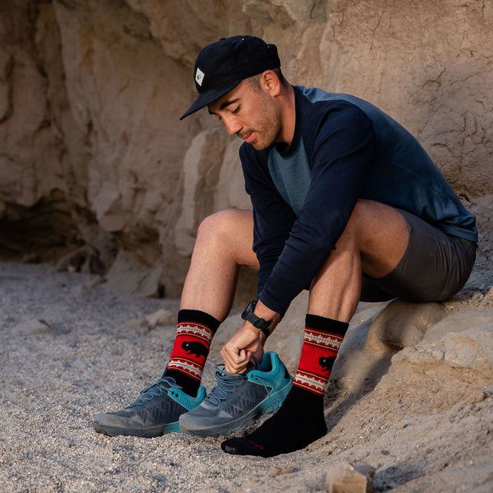 Shot of model sitting on a rock wearing men's vangrizzle boot hiking sock in black while putting on blue hiking shoes