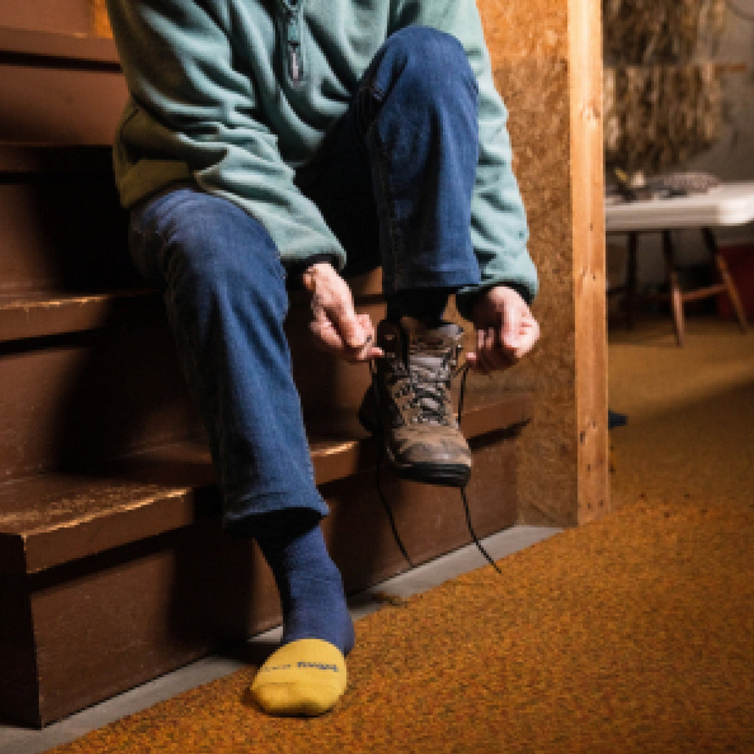 Model sitting on wooden stairs inside wearing 2015 socks in indigo and pulling on brown work boot on left foot