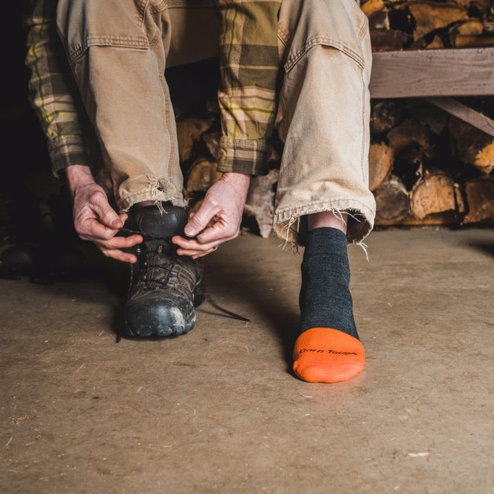 Model sitting on bench in front of wood pile wearing 2017 socks in graphite colorway and lacing up brown work boot on right foot.