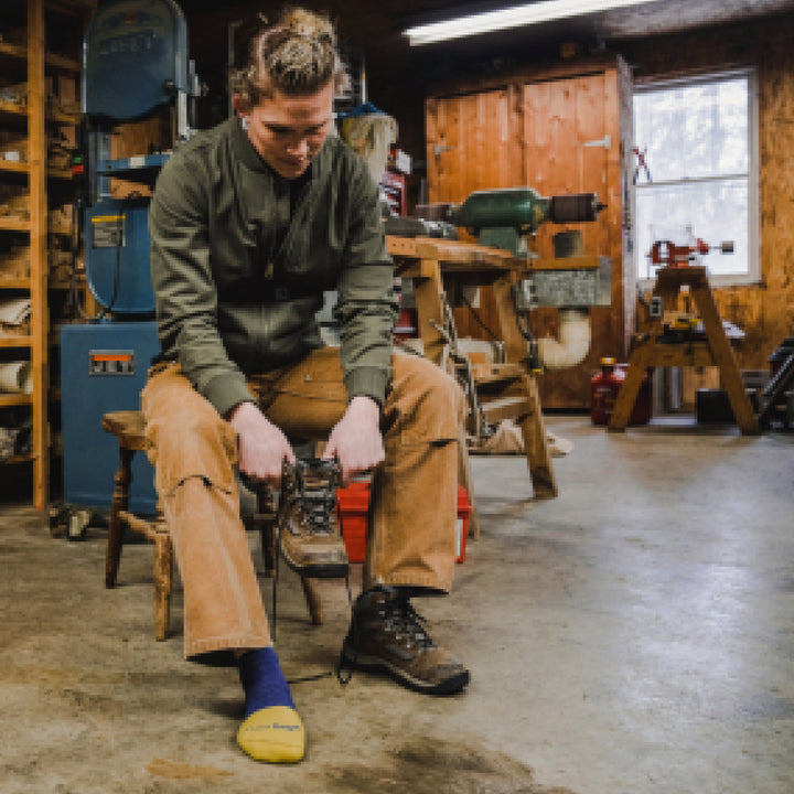 Model sitting on stool in a shop wearing 2204 socks in Indigo colorway with brown boot on left foot and holding the other