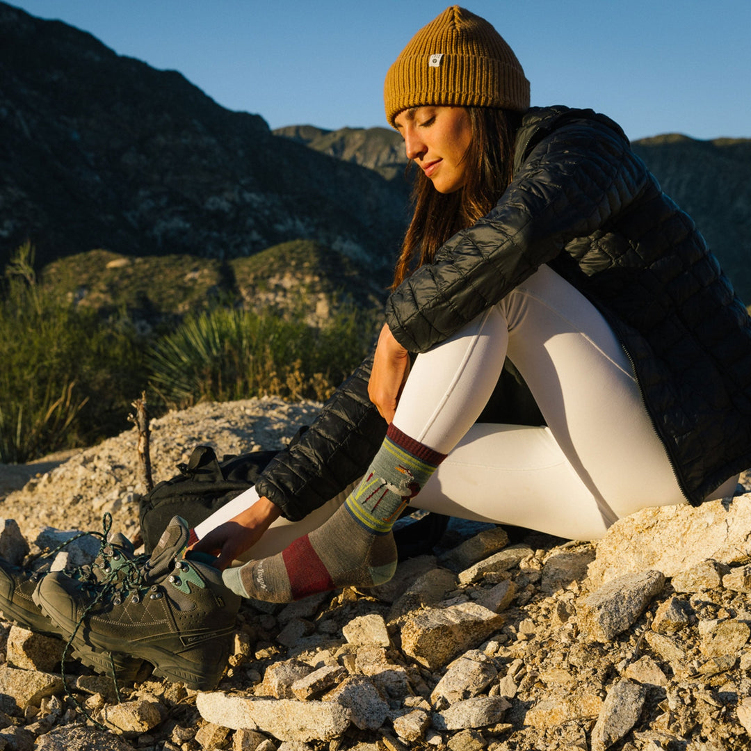Model sittting down wearing women's critter club micro crew lifeestyle sock in taupe with mountains in background
