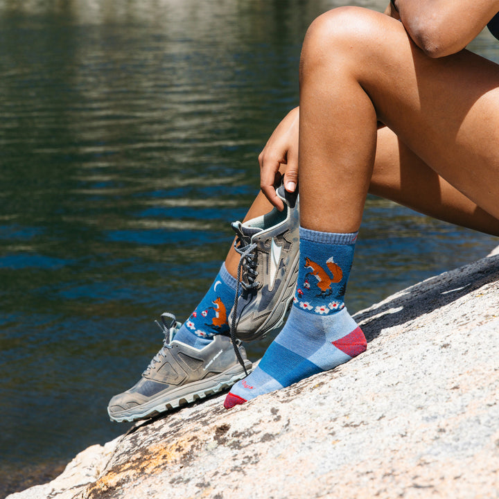 Close up shot of model sitting next to a lake wearing the women's critter club micro crew hiking sock in vapor blue