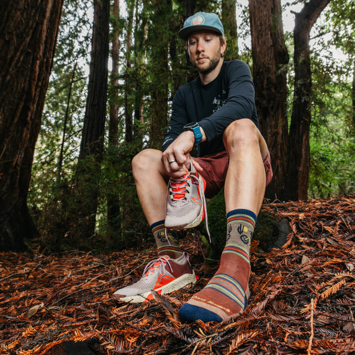 Model sitting on a rock in the woods putting on sneaker wearing the 5003 Willoughby Micro Crew Hiking Sock in Chestnut