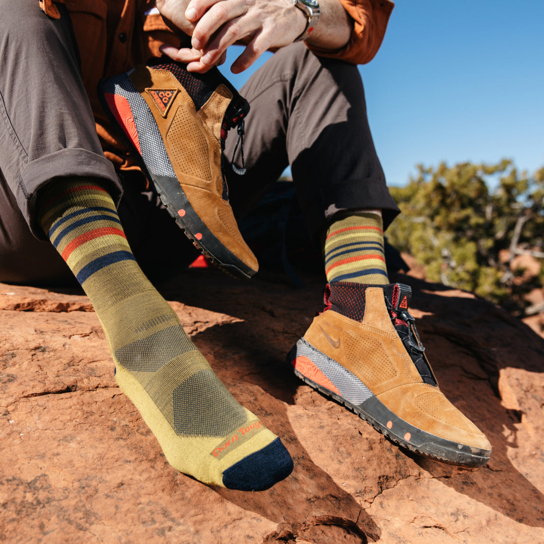 Image of model sitting on red desert rocks wearing 5012 socks in Sandstone colorway while wearing one brown hiking boot and holding the other