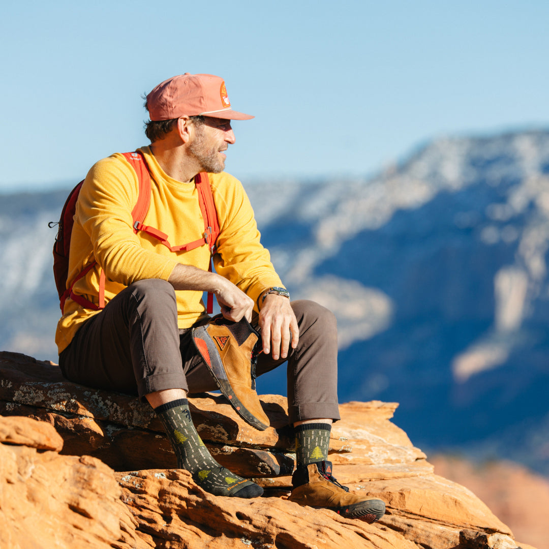 Image of model sitting on red desert rocks wearing 5015 socks in forest colorway while wearing one brown hiking boot and holding the other