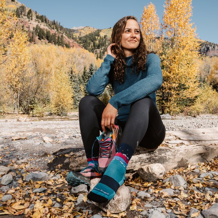 model sitting next to riverbed one shoe off show the 5025 mirror lake micro crew hiking sock in teal colorway