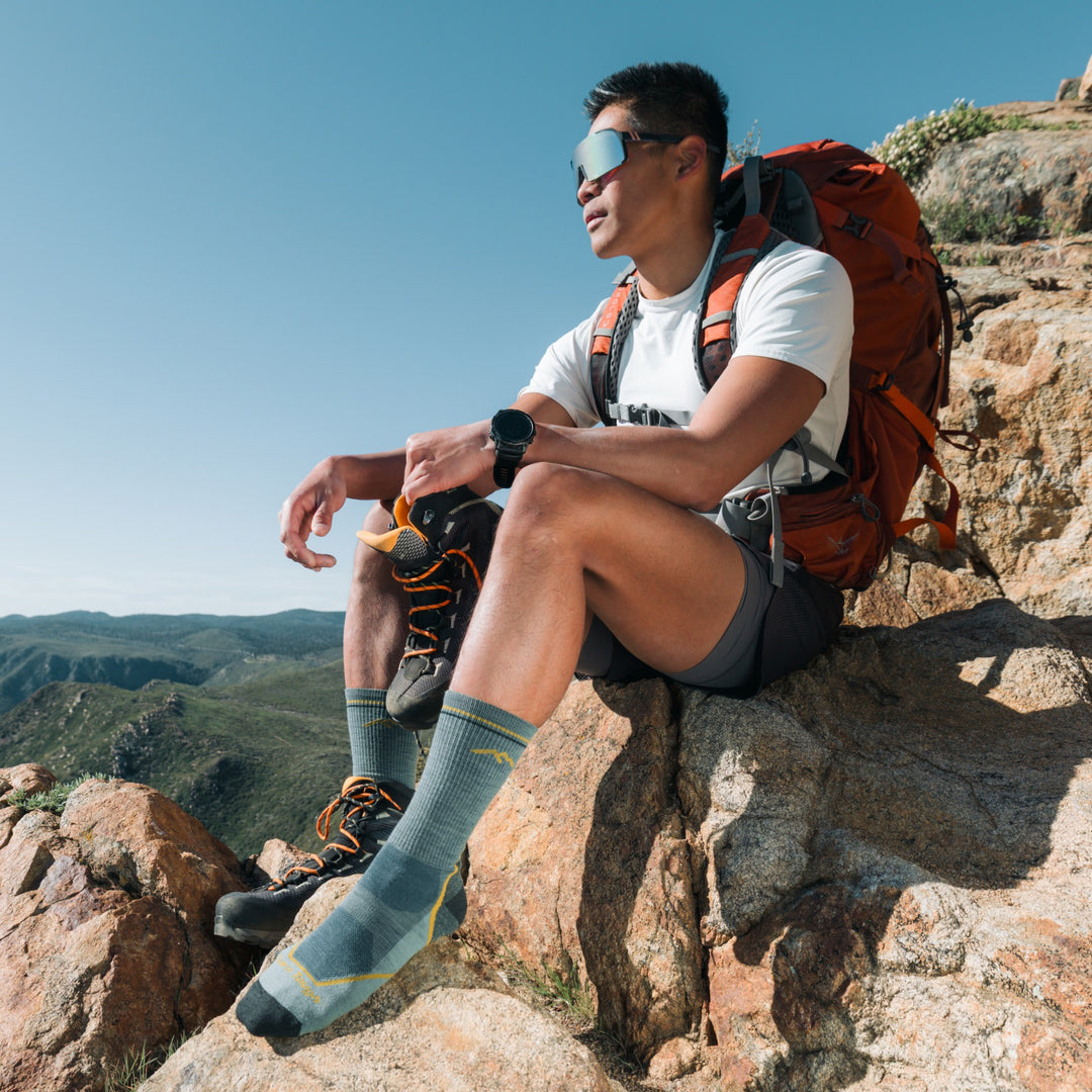 model sitting on rock ledge holding one shoe wearing the 5028 light hiker boot sock in seafoam green colorway
