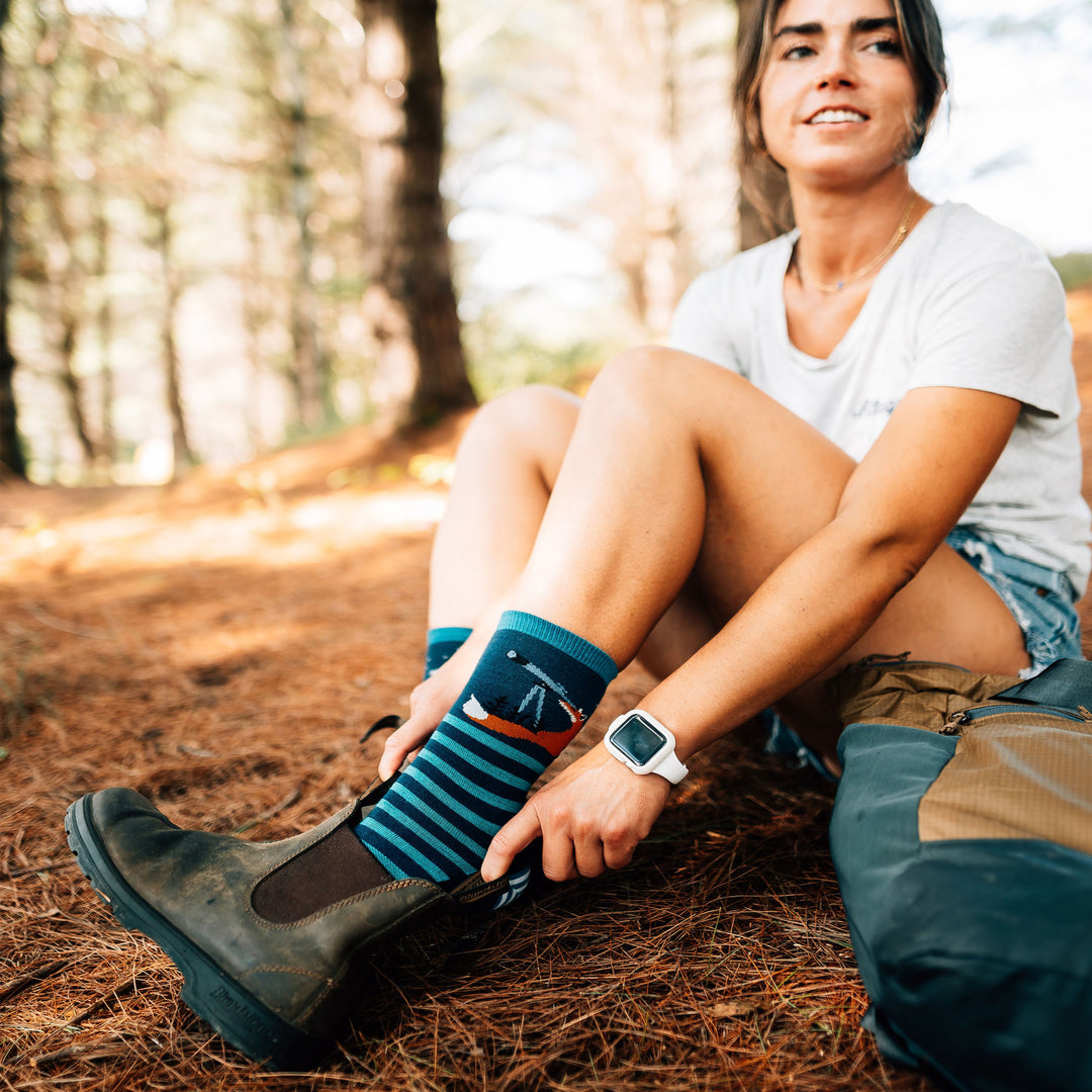 Model sitting in the woods putting on boots wearing the women's animal haus crew lifestyle sock in eclipse