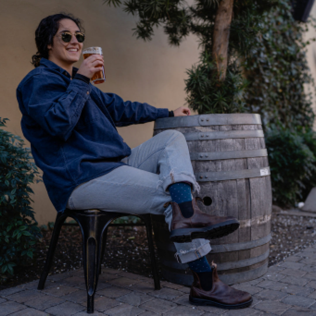 Model sitting next to barrel while drinking beer wearing brown boots and 6110 socks in Dark Teal colorway 