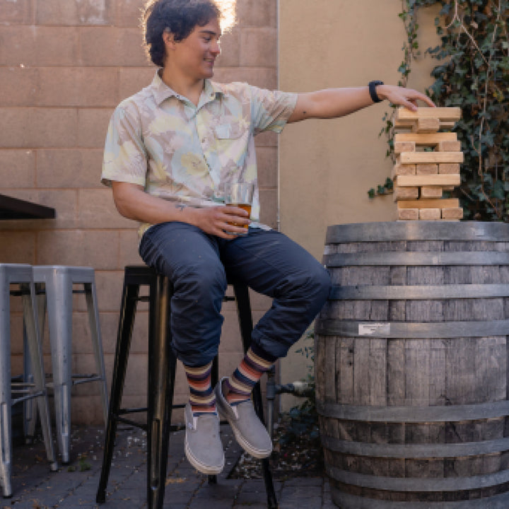 Model sitting on stool holing glass of beer while playing wood block game and wearing 6113 socks in Bark colorway with gray sneakers