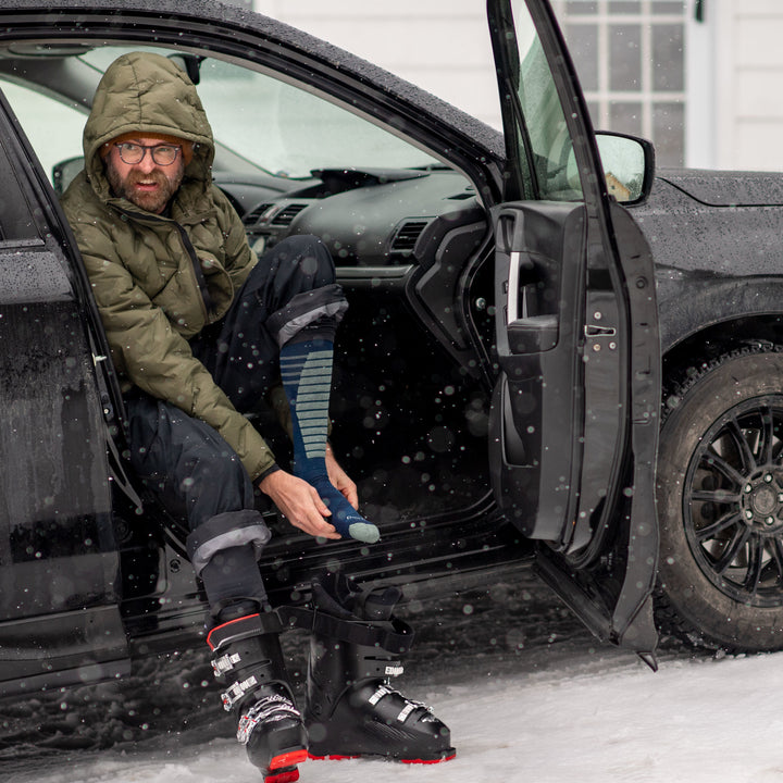 Model sitting in a car wearing men's edge over-the-calf snow sock in midnight blue while putting on black ski boots