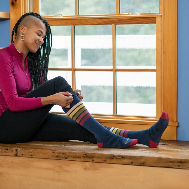 Full shot of model sitting on a wood window sill putting on women's snowburst over-the-calf snow sock in denim blue