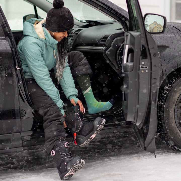 Full shot of model sitting in a car wearing women's aurora over-the-calf snow sock in aqua while putting on black ski boots