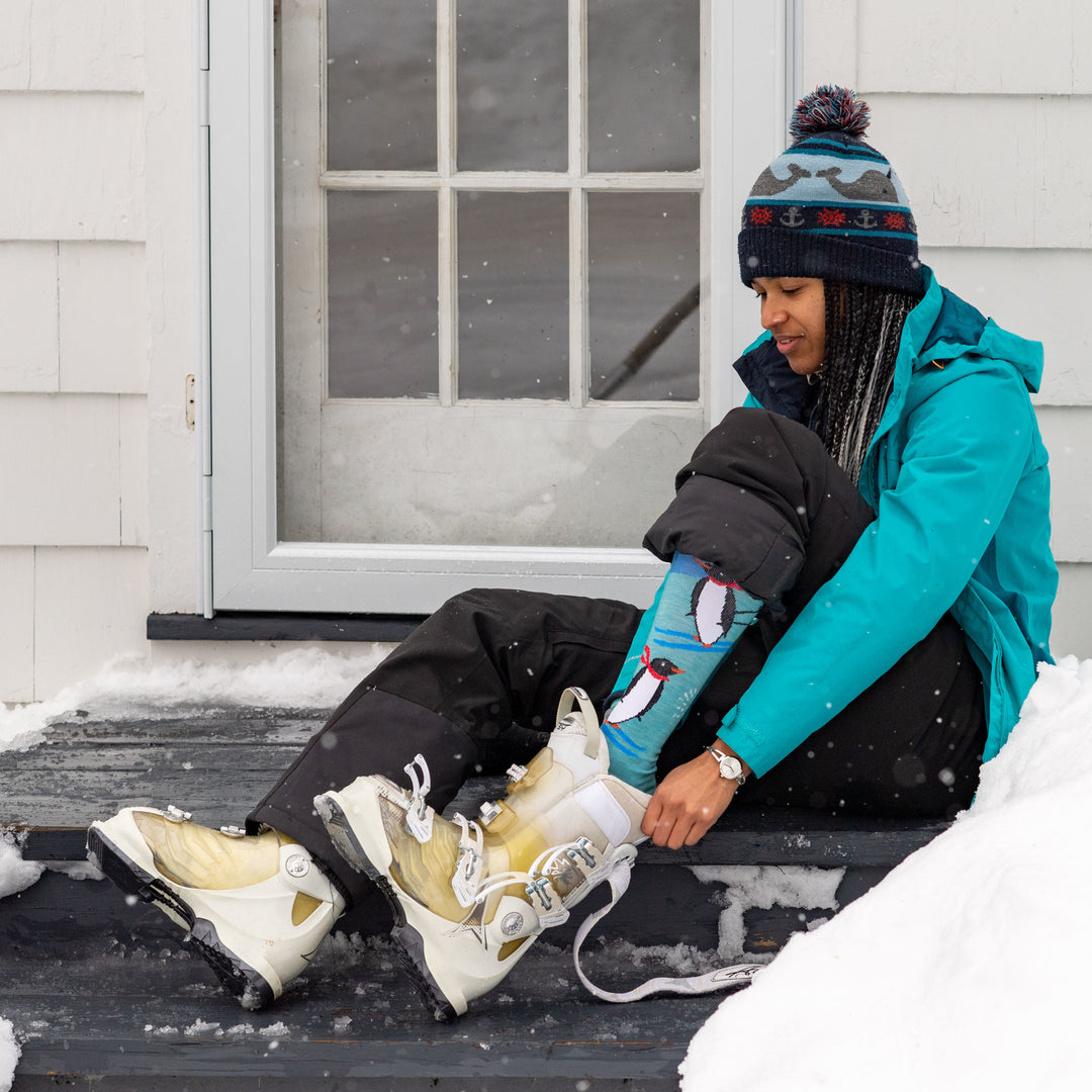 Model sitting outside a house wearing women's penguin peak over-the-calf snow sock while putting on white ski boots
