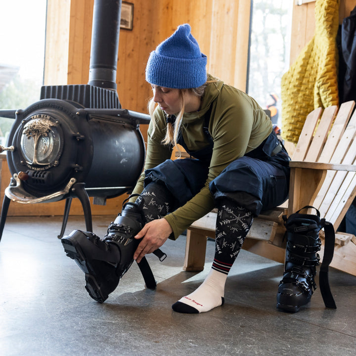 model sitting on edge of chair next to woodstove putting on ski boots wearing 8060 snowfall OTC midweight with cushion sock black