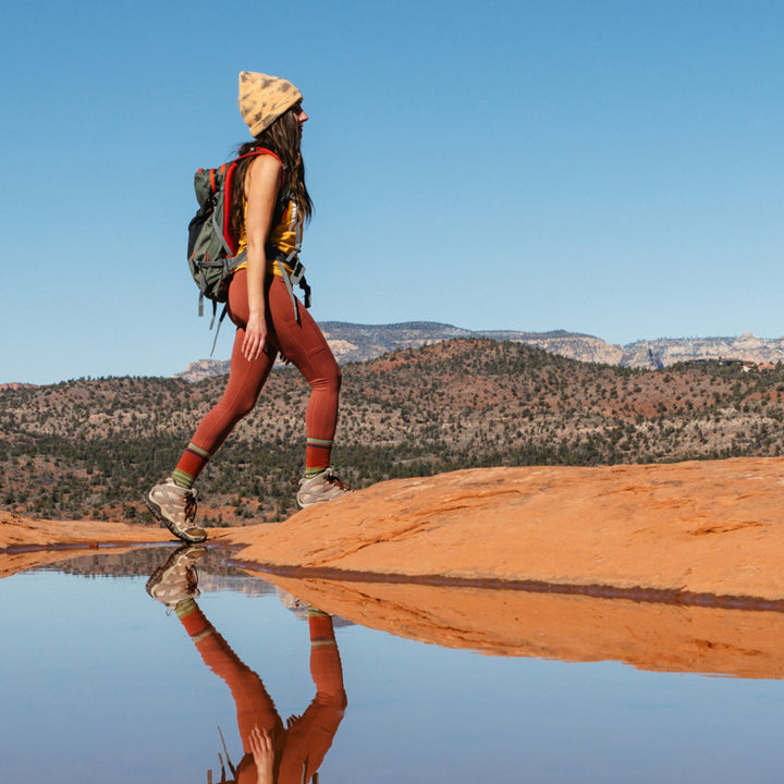 Wide shot of model hiking across red desert rock wearing 1969 socks in sandstone colorway with figure reflected in a pool of water in the foreground and desert mountains in the background
