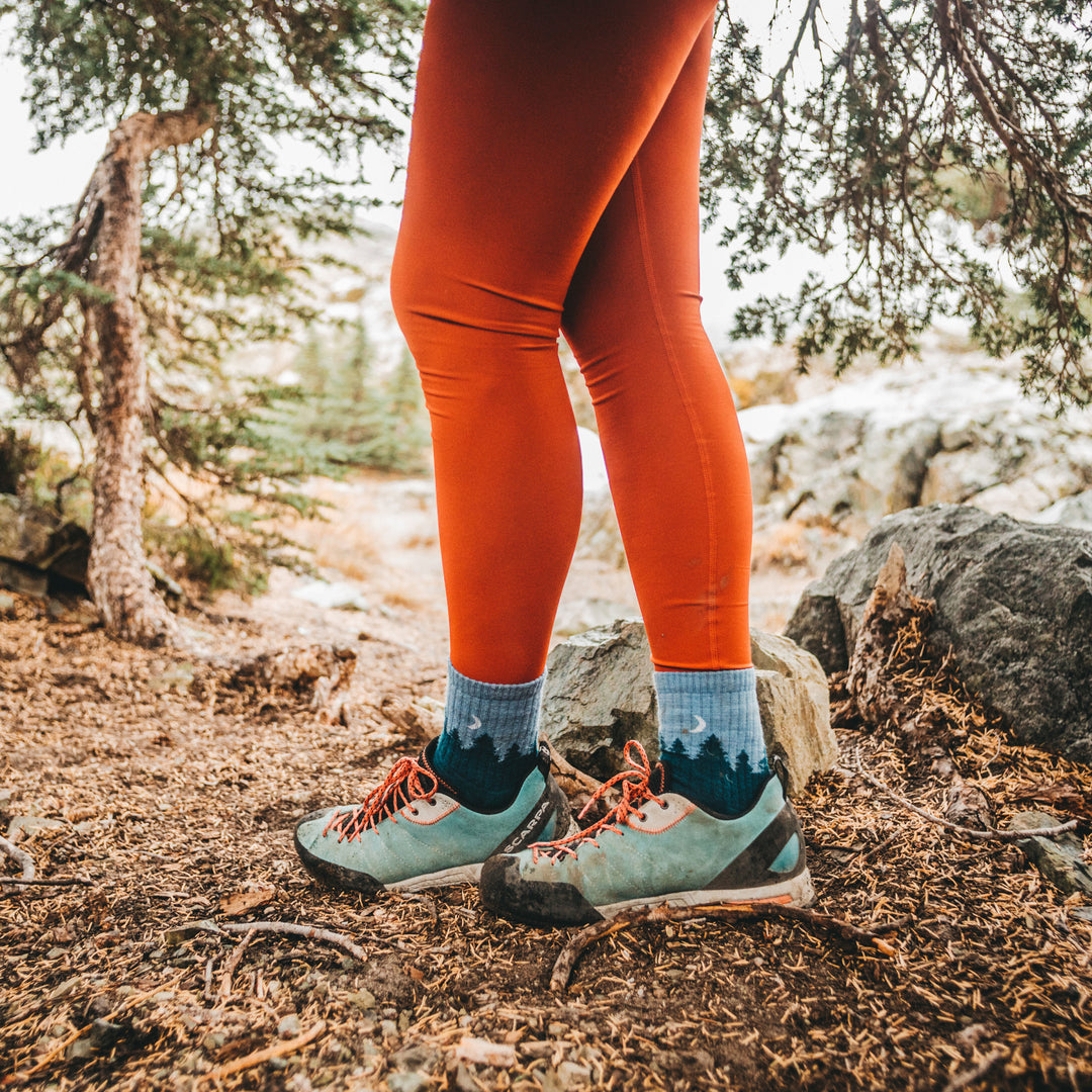 Female hiker wearing orange leggings in a forest, feet crossed at the ankle wearing womens treeline micro crew hiking socks in blue, Lifestyle Image