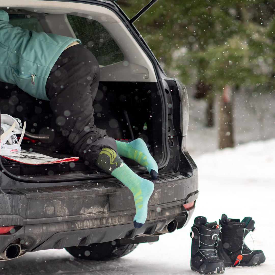 Model in trunk of a black car with her feet hanging out wearing women's aurora over-the-calf snow sock in aqua