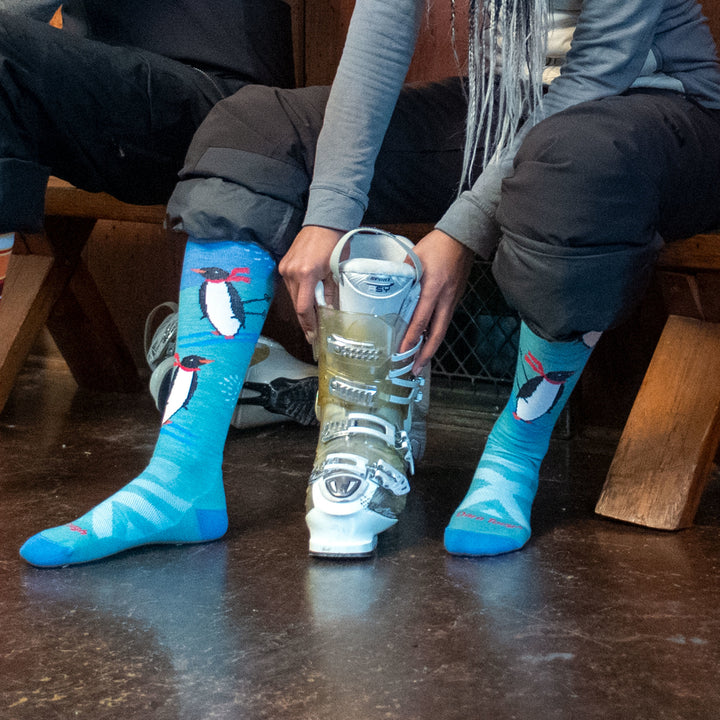 Shot of model sitting down wearing women's penguin peak over-the-calf snow sock while putting on silver ski boots