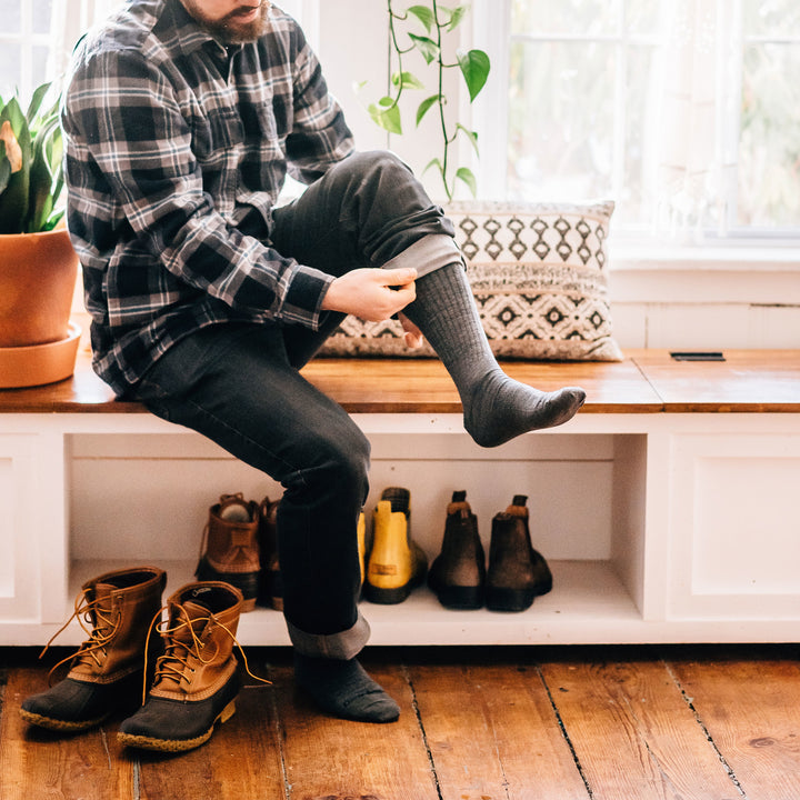 Man sitting on a wooden bench in his house wearing The Standard Mid-Calf Lightweight Lifestyle Sock in Medium Gray