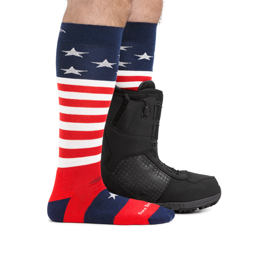 Man standing facing right, front foot barefoot wearing Captain Stripe Over the Calf Ski & Snowboard Socks in Stars and Stripes pattern and back foot wearing the sock and a snowboard boot, showing the height of the sock comes to just below the knee