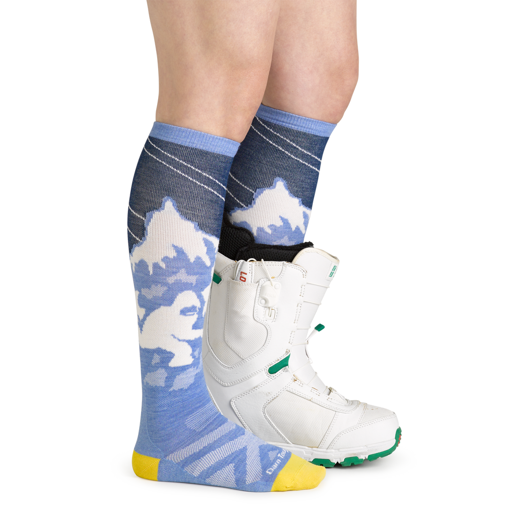 Studio shot of model wearing women's yeti over-the-calf snow sock in midnight with white snowboard boot on left foot