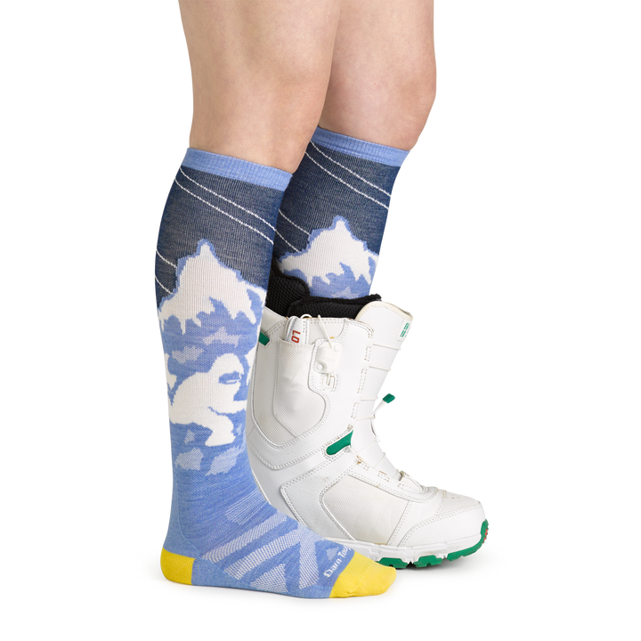 Studio shot of model wearing women's yeti over-the-calf snow sock in midnight with white snowboard boot on left foot