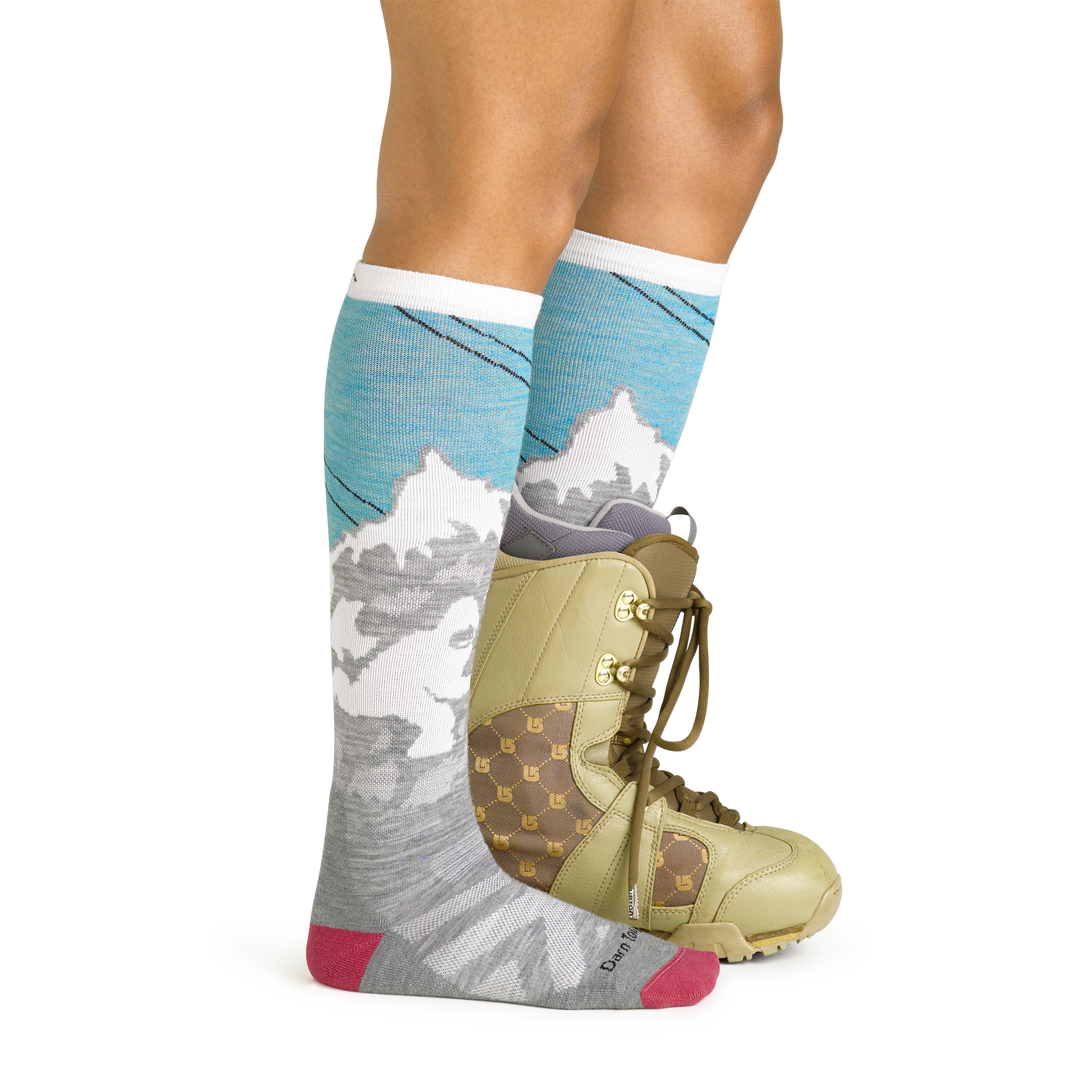 Side studio shot of model wearing women's yeti over-the-calf snow sock in aqua with green snowboard boot on left foot