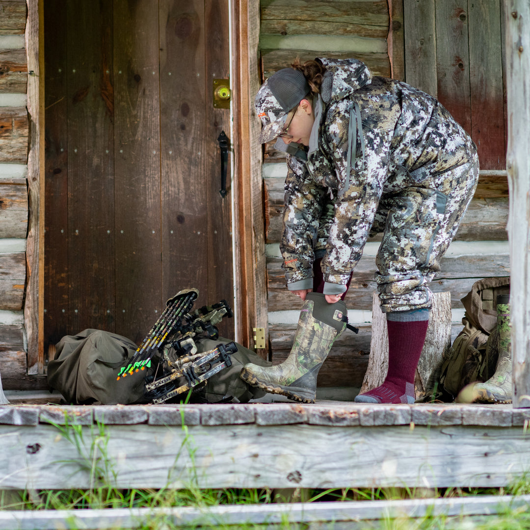 Lifestyle shot of woman in hunting gear on her cabin porch. She is putting her Muck Boots on over her hunter socks.