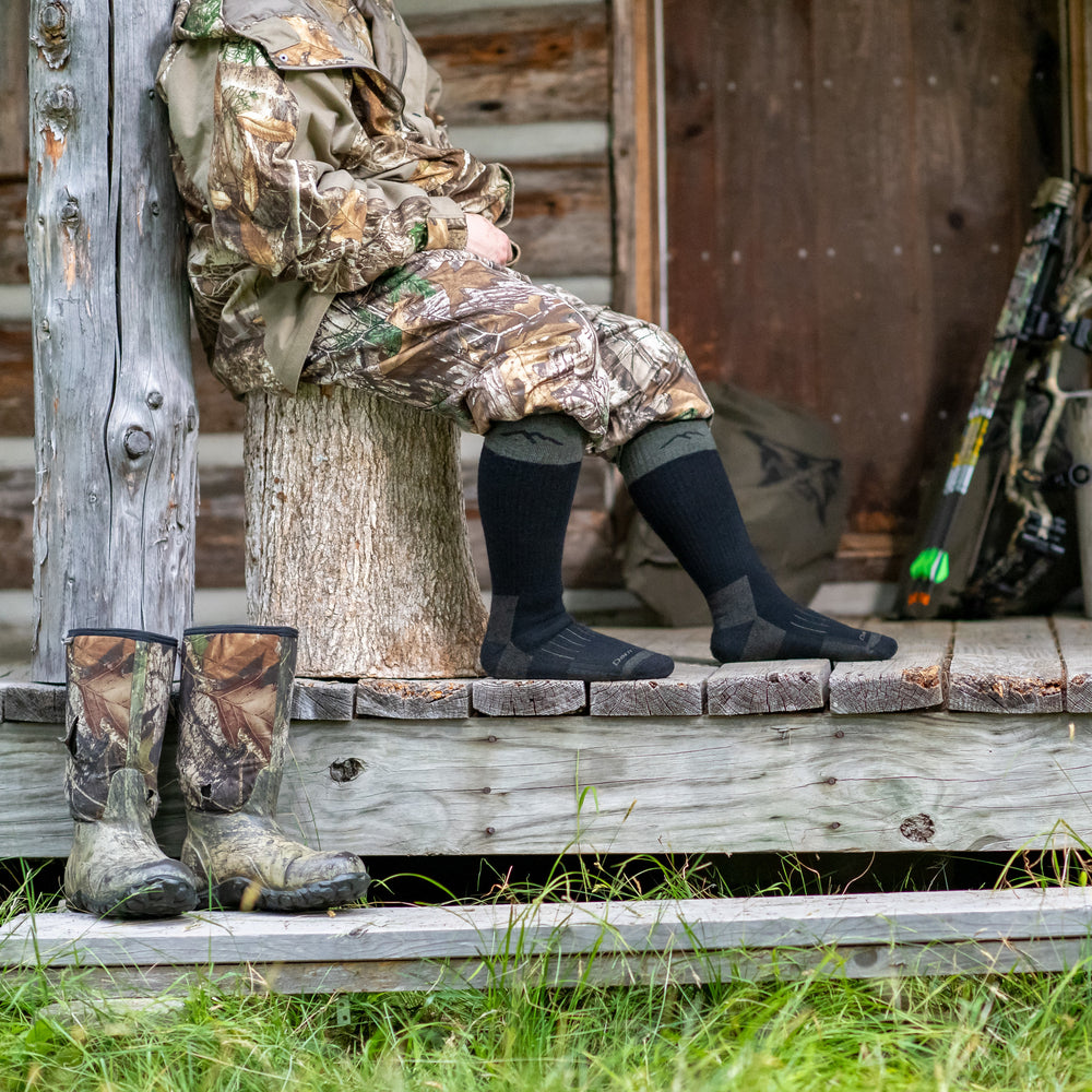 Side shot of model sitting on a tree stump in camouflage wearing the unisex boot hunting sock in charcoal