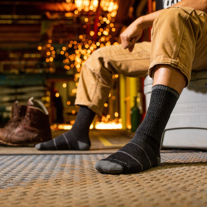 Side shot of model sitting on a cooler in a workshop wearing the men's william jarvis boot work sock in gravel