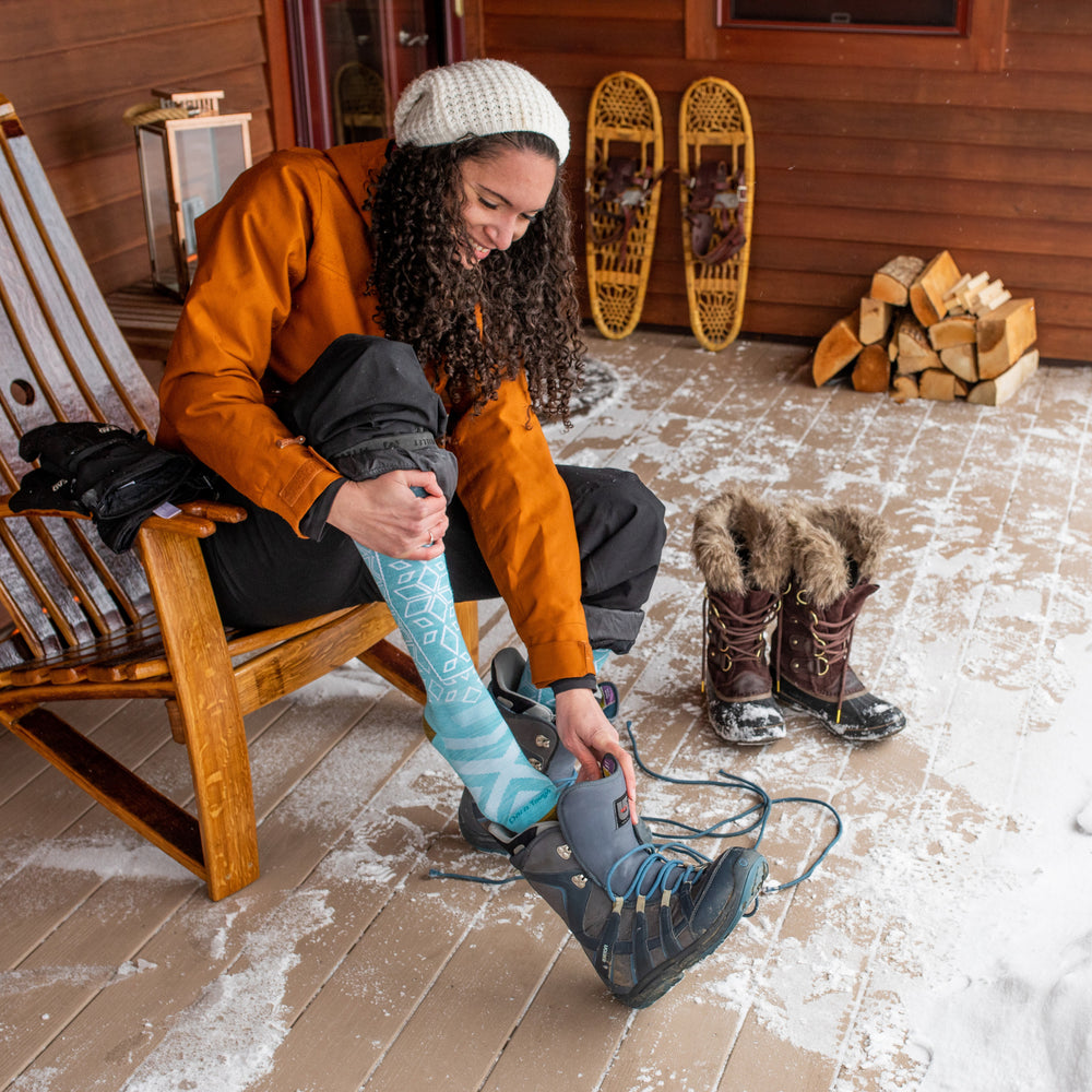 Lifestyle shot of woman lacing up her snowboard boots in Adirondack chair on her cabin porch wearing Traverse socks in Aqua.