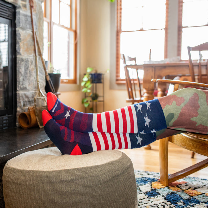 Side shot of model sitting by a fireplace with their feet up wearing the men's captain stripe over-the-calf snow sock