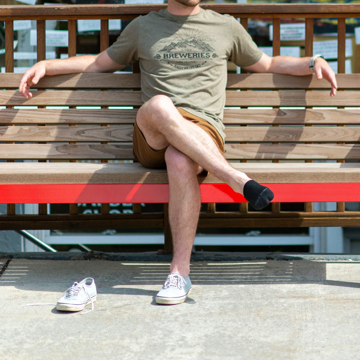 Full shot of model sitting on a park bench with one white shoe on wearing the men's   no show hidden lifestyle sock in black