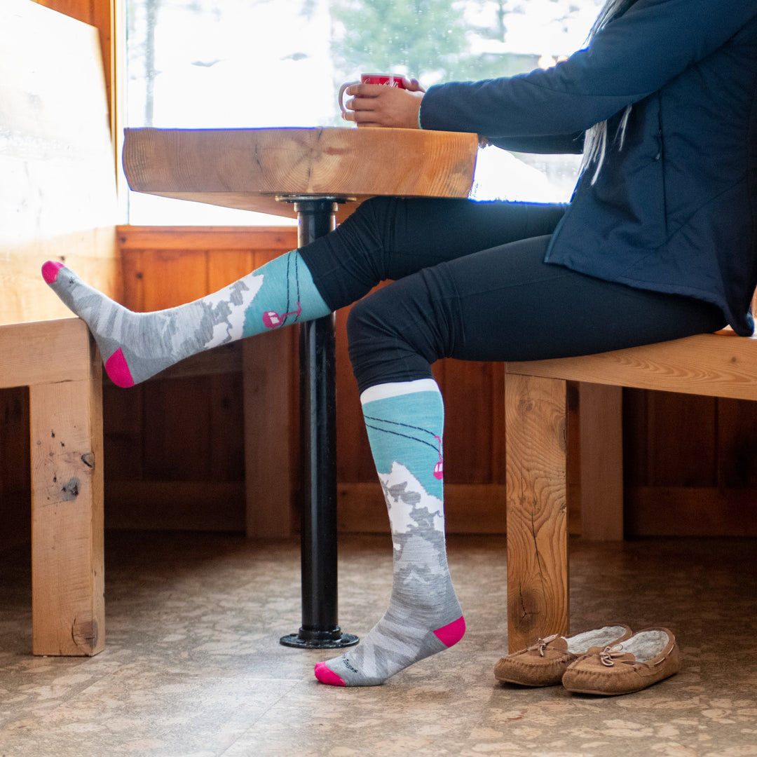 Lifestyle image of woman sitting at café table with a cup of coffee, getting ready to hit the slopes in her Yeti socks.