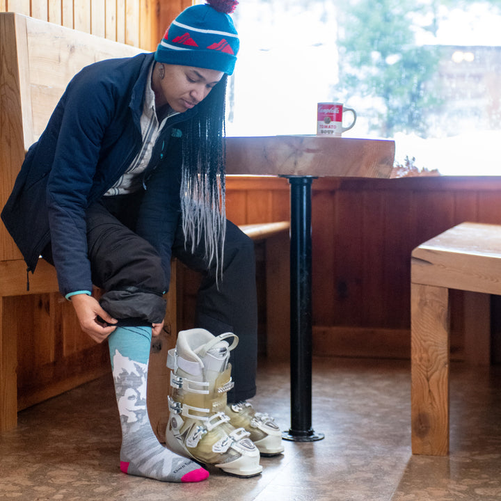Lifestyle image of woman sitting at café table, buckling up her ski boots in her Yeti socks.