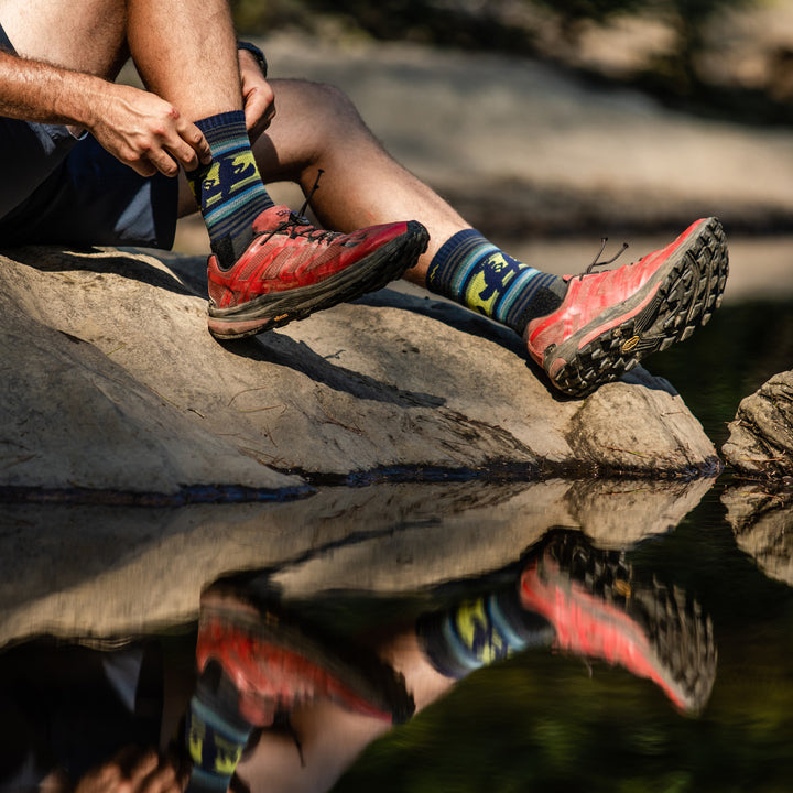 Close up shot of model sitting on a rock by the water putting on the men's willoughby micro crew hiking sock in forest